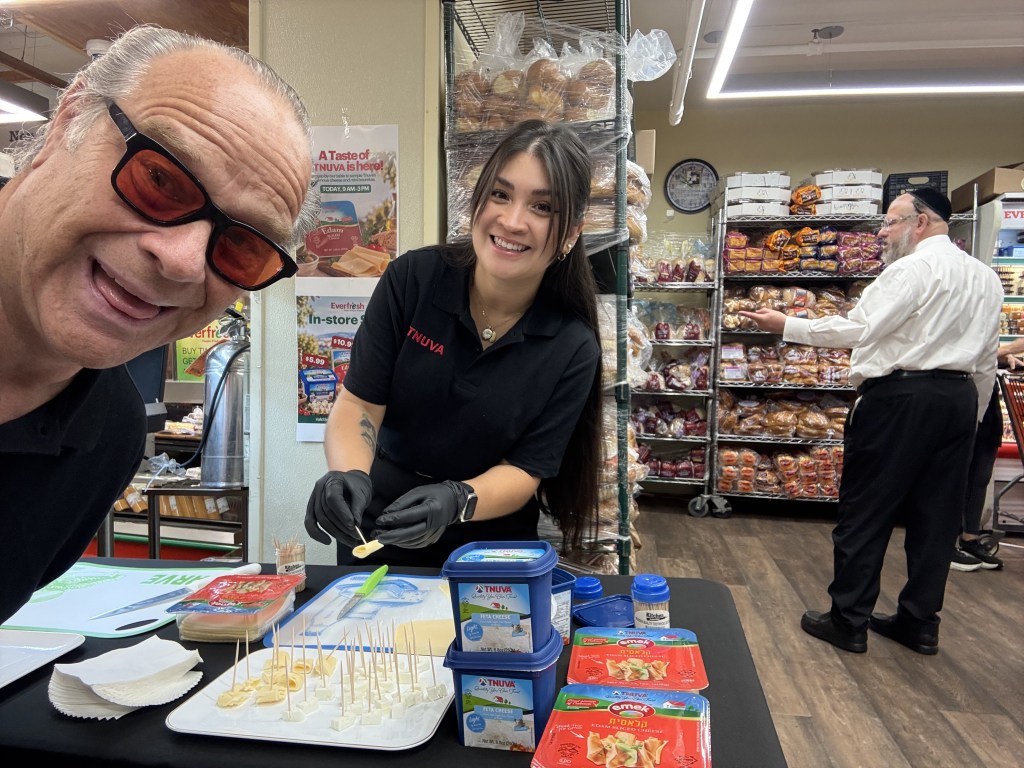 A man and a woman smiling at a food sampling event. The woman, wearing a black T-shirt with 'TNUVA' on it, is preparing food samples on a table covered with various containers and food items. In the background, a man in a white shirt is observing the product display in a grocery store.